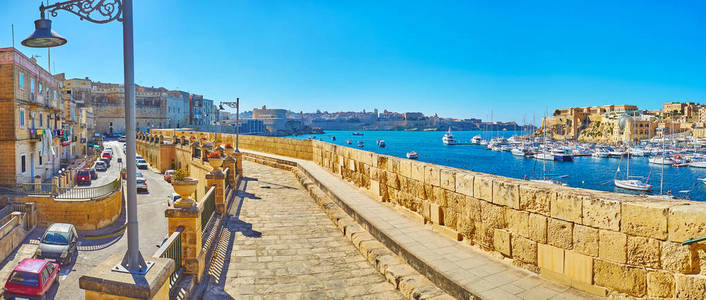 Birgu 海滨长廊全景, 沿着城墙伸展, 以老式的路灯, 在 Kalkara 码头的船只和在背景上的瓦莱塔地平线, 马耳他