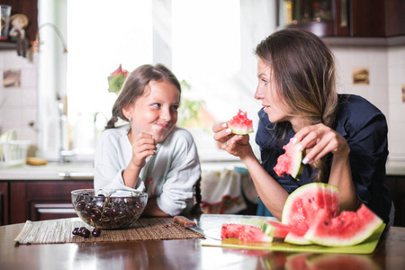 可爱的小女孩和她美丽的妈妈正在切水果, 红色西瓜和微笑, 而在厨房做饭在家里。幸福的家庭