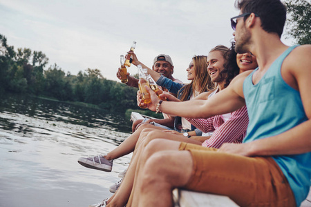 快乐的年轻多民族的朋友在休闲装微笑和烘烤啤酒瓶, 而坐在河码头上