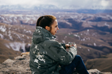 一个穿着暖和夹克的年轻女孩坐在悬崖边上, 享受着高山的自然和清新的空气。