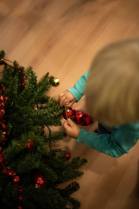 在室内圣诞树上悬挂红色节日球的金发小男孩的最高视图