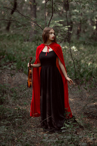 优雅的神秘女孩黑色礼服和红色斗篷在黑暗的树林里握小提琴