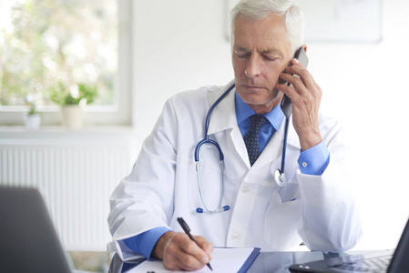 男性医生肖像打电话与他的病人和写下诊断坐在咨询室