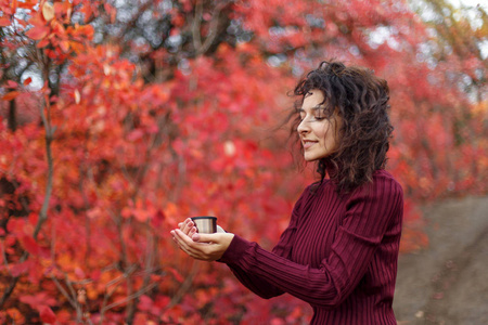 年轻的 blackhaired 妇女在红色 sweather 与杯子热的饮料在红色 autumnn 灌木