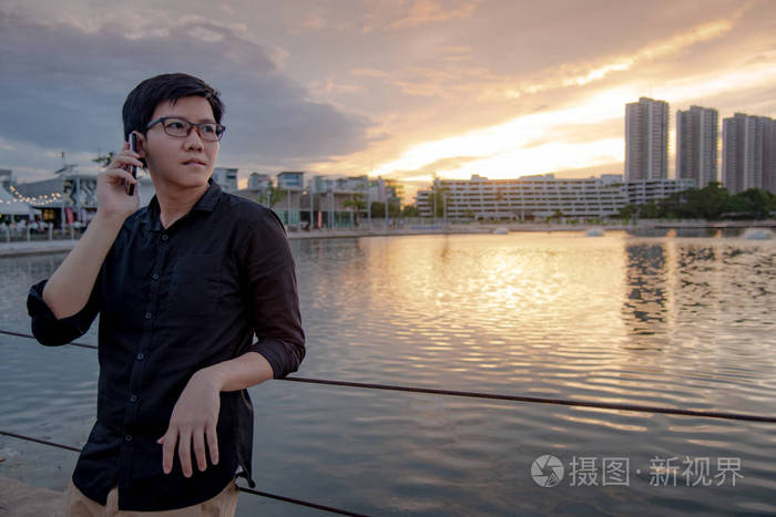 亚洲人与眼镜使用智能手机在城市的湖泊在日落期间。手机上的商务沟通。城市生活方式概念
