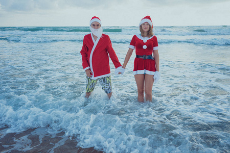 快乐的情侣在海滩上漫步圣诞老人的服装。圣诞节假期的概念