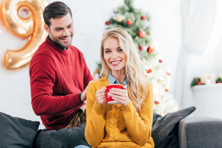 美丽的快乐的女人拿着咖啡杯, 而男人看着她在家里与圣诞树