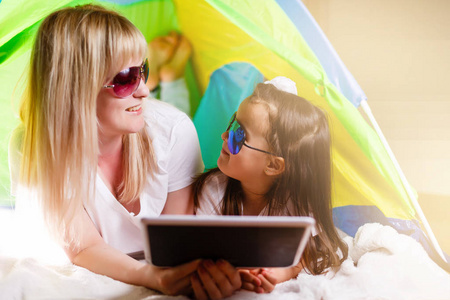 快乐的母亲和她的小女儿戴着墨镜用平板电脑躺在帐篷里