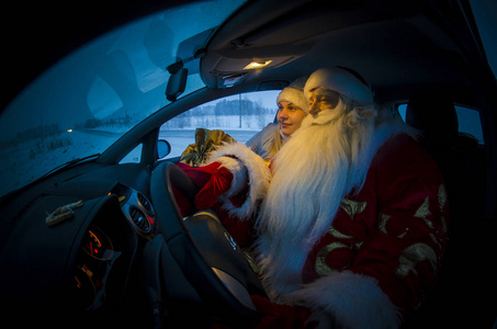 滑稽的圣诞老人和他的美丽的孙女坐在汽车在晚上和问候