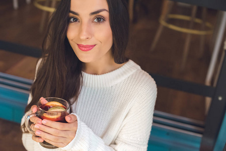微笑的年轻妇女看着照相机和拿着杯子在咖啡桌上的酒
