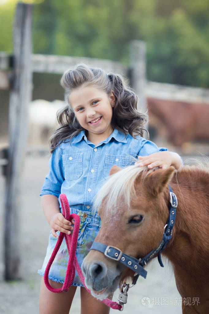 一个小的微笑的女孩卷曲的头发穿着牛仔裤与小马在稳定