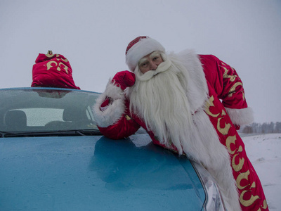 圣诞老人站在蓝色的汽车与一袋礼物和挥手祝贺