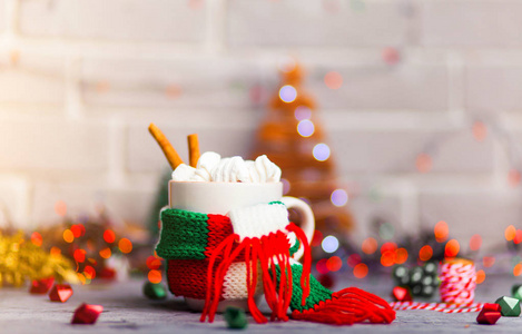 杯热咖啡或茶与迷你棉花糖和肉桂与绿色的红色白色围巾对一个乡村背景与美丽的圣诞灯散景玩具