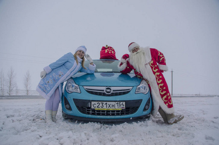 滑稽的圣诞老人和他的美丽的孙女微笑和站立在蓝色汽车附近与一袋礼物和祝贺