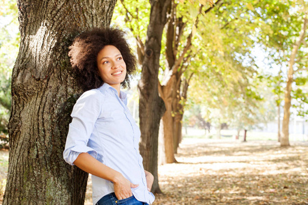 户外肖像的快乐美丽的非洲裔美国妇女在自然界中, 女性在蓝色的衬衫享受夏日, 靠在公园的树上, 看着侧面