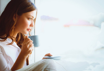 年轻女子在家里坐在靠近窗口轻松的在她的客厅里看书 喝咖啡或茶