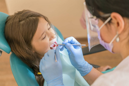 可爱的小女孩躺在牙医的椅子上, 微笑着在相机上, 而口腔检查由牙医进行使用探针。坐在牙医办公室的小女孩
