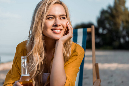 有吸引力的微笑的女孩坐在太阳躺椅与啤酒在瓶里和看离开