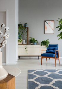 图案地毯和蓝色扶手椅在灰色客厅内部与海报和植物。真实照片
