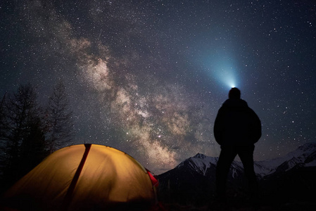 男子游客与手电筒站在他的帐篷附近, 晚上在一个充满星星的天空下