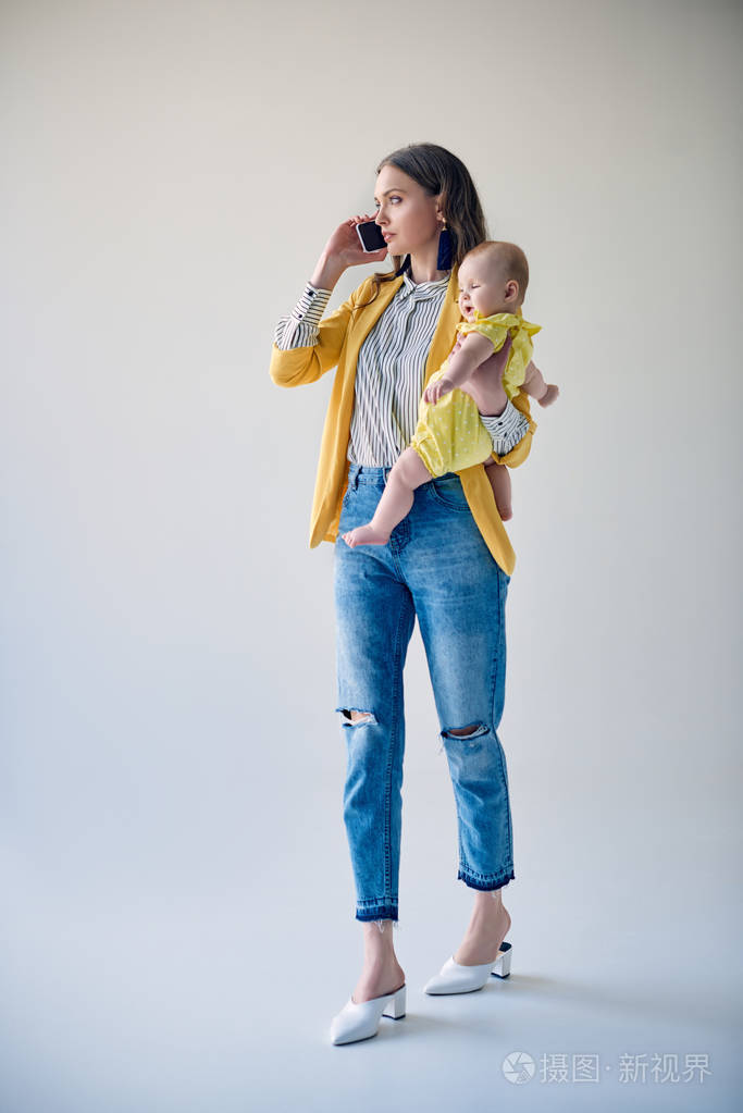 时尚的母亲携带可爱的婴儿女儿和智能手机交谈灰色