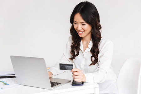 微笑着年轻的亚洲女商人手持塑料信用卡, 而坐在笔记本电脑以上的白色背景
