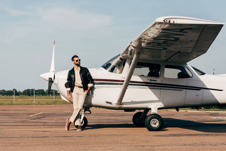 年轻的时尚男性飞行员在皮革夹克和太阳镜摆在飞机附近