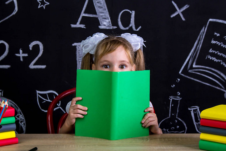 女生坐在书桌前, 脸上有一本书, 藏在书后, 四周都是学校用品。黑板作为背景。放大视图