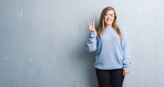 年轻的成年妇女在灰色的难看的垃圾墙戴眼镜显示和指向与手指数字三同时微笑自信和快乐