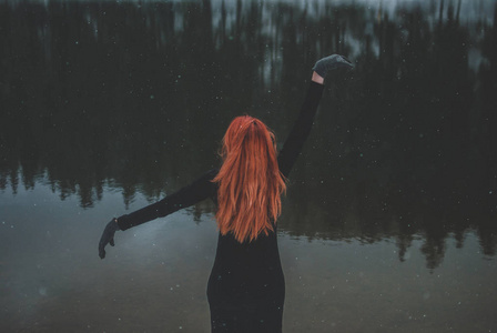 雪花飞过英俊的红发女孩看着远处, 举起双手在她的头顶上。灿烂阴暗的黑山湖。从后面看