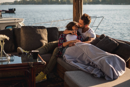 男友亲吻他的女友在额头, 而她躺在河边覆盖着毯子。美丽的秋日
