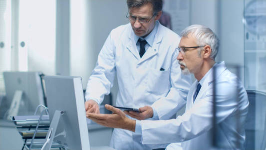 两名资深医生在桌面计算机讨论医疗问题。其中之一举行的平板电脑。办公室是现代和光明