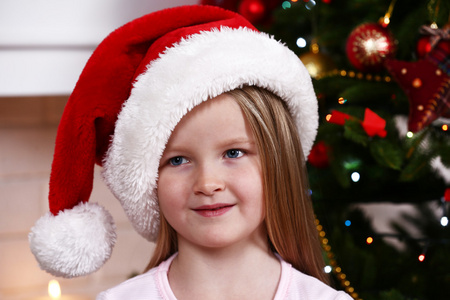 在壁炉背景下靠近杉树的圣诞老人帽的小女孩
