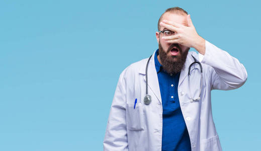 年轻的高加索医生在孤立的背景上穿着医学白色外套, 用手在震惊的脸上偷看, 用令人尴尬的表情通过手指看着