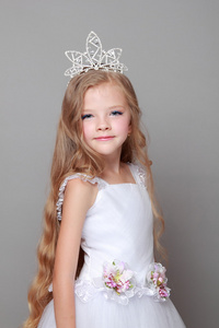 美丽的长头发在冠和可爱的笑容摆在美丽和时尚的灰色背景上的摄像头白色礼服的白人小女孩