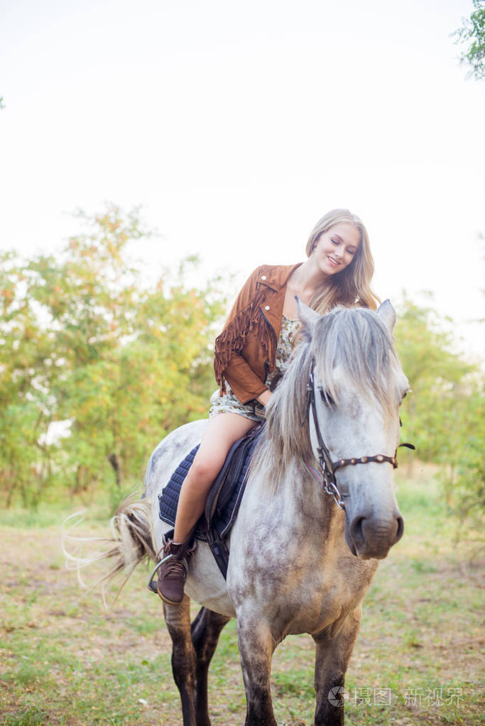 美丽的年轻女孩长着金色的头发在一个麂皮绒夹克与条纹微笑骑着马在