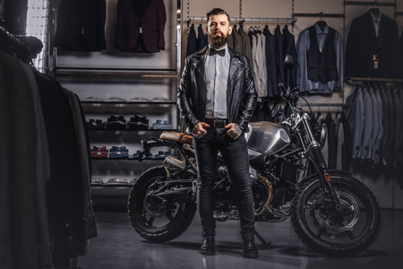时尚纹身胡子男子身着黑色皮革夹克和弓领带摆在男装服装店附近复古运动摩托车