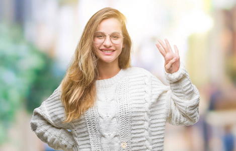 年轻美丽的金发女子穿着冬季毛衣和太阳镜在孤立的背景显示和指向与手指数字四同时微笑自信和快乐