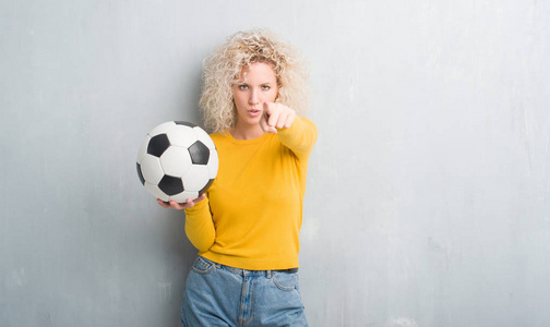 年轻的金发碧眼的女人在格格灰色背景举行足球足球用手指指向相机和你, 手的标志, 积极和自信的手势从前面