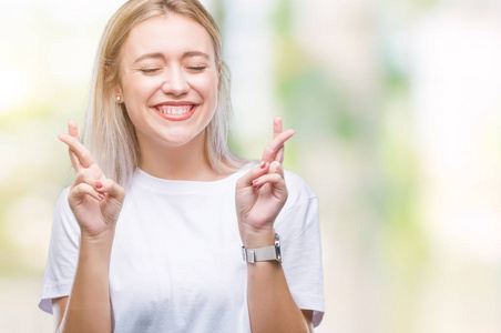 年轻的金发女人在孤立的背景下微笑着交叉手指, 希望和眼睛闭上。运气和迷信概念