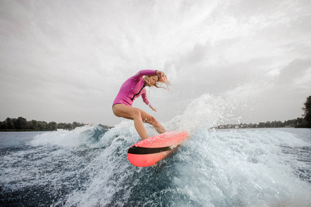 活跃的金发女子 wakesurfer 身着粉红色泳装骑在阴天的蓝色飞溅波