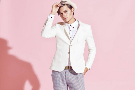 肖像的年轻 英俊 严重 现代 男模穿着白色夹克 白衬衫 灰色裤子和白色帽子摆在浅粉红色背景的工作室