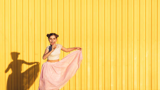 快乐时尚的女孩在粉红色的裙子沾了她的鼻子与冰淇淋和微笑对黄色墙壁
