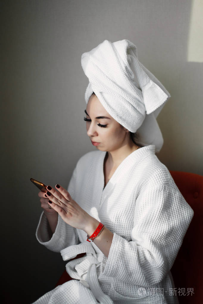 一个年轻的女孩淋浴后, 一个智能手机裹在毛巾坐在窗边的椅子上