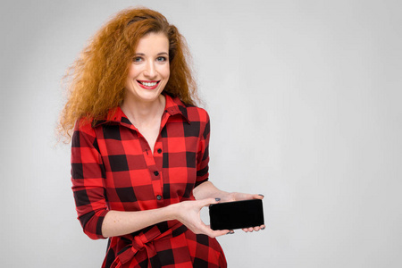美丽的卷发红头发的白种女人在红色格子礼服显示不同的情感表现在白色墙壁在演播室。微笑和显示空白智能手机