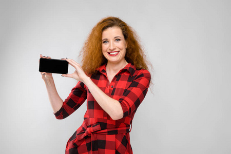 美丽的卷发红头发的白种女人在红色格子礼服显示不同的情感表现在白色墙壁在演播室。微笑和显示空白智能手机