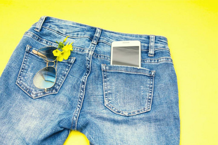 蓝色牛仔裤口袋里的太阳镜手机和黄色花朵