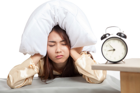 困倦的亚洲女孩用枕头捂住耳朵和闹钟照片