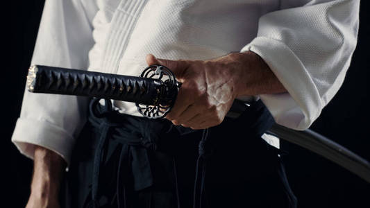 合气道大师穿传统武士袴衣服持有日本剑入鞘。他是在聚光灯下黑暗包围他。孤立在黑色背景上的镜头