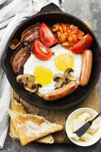 传统的英国早餐培根, 香肠, 煎蛋, 西红柿, 豆类, 蘑菇, 烤面包用 butterin 烹饪锅和咖啡的灰色背景。顶部视图